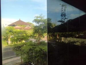 Perbandingan kaca biasa dan kaca film gedung di Bali — sisi kiri tanpa film terlihat tembus pandang, sisi kanan dengan kaca film gelap memantulkan pemandangan pura dan pepohonan khas Bali