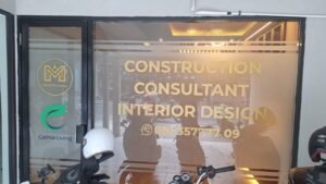 Cutting sticker emas M Construction dan logo Calma Living pada kaca depan kantor